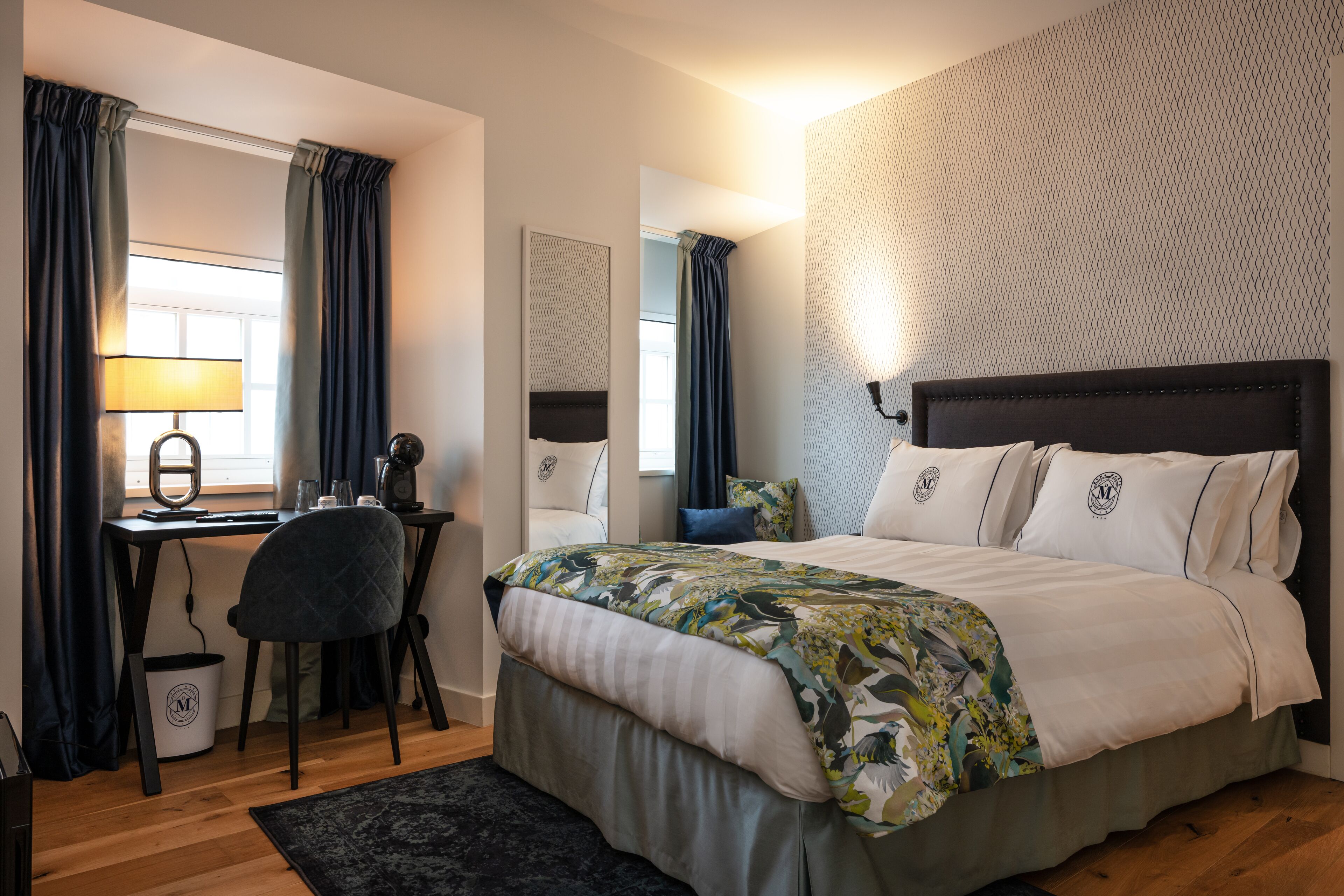 Quarto de hotel moderno com cama, escrivaninha, cadeira e uma janela grande com cortinas.