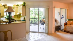 A room with a kitchen area, a dining table with chairs, a bed, and a sliding glass door leading to an outdoor area with a chair and plants. - Jupiter Bay Beach Condo (Jupiter)