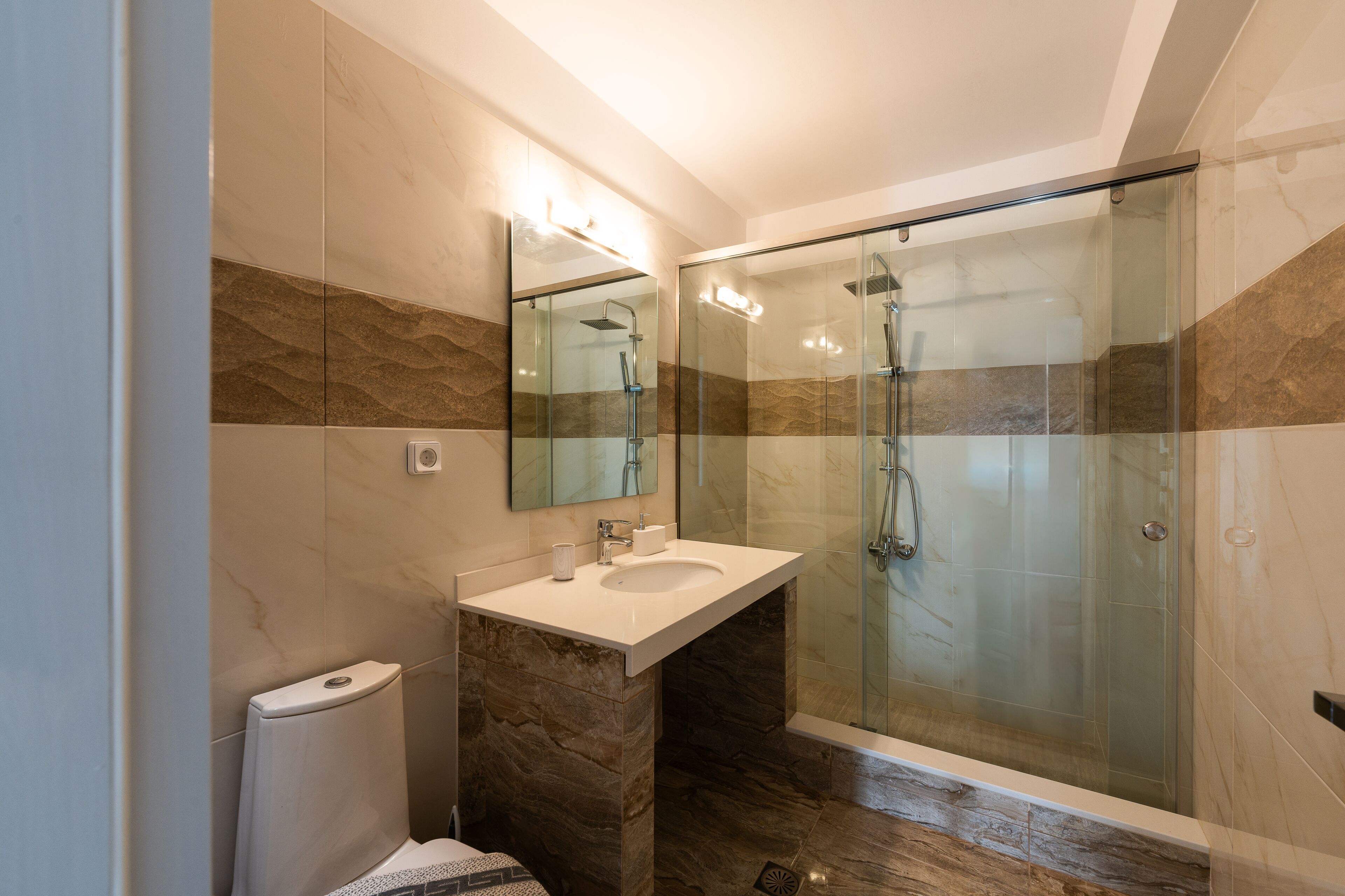 Un bany modern amb dutxa envidrada, paret revestida de marbre i un lavabo blanc.