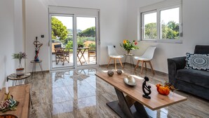 A spacious living room with a large sliding glass door leading to an outdoor patio. There is a wooden coffee table with decorative items, a black leather sofa, and a white chair. - Sweet Home Apartments (Rhodes)