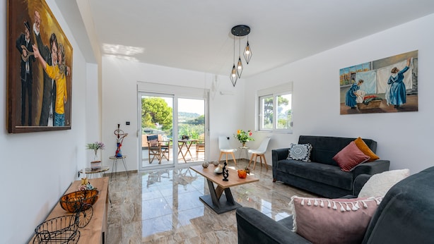 A spacious living room with a large window, a black sofa, a wooden coffee table, and a painting on the wall. - Sweet Home Apartments (Rhodes)