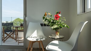 A modern room with a large window, two white chairs, a small table, and a potted plant with red flowers. - Sweet Home Apartments (Rhodes)