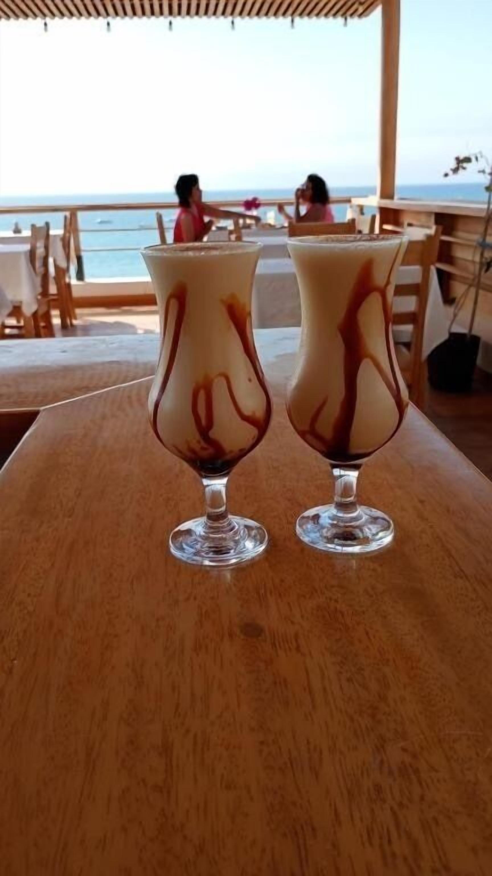Two tall glasses of a layered beverage on a wooden table at a seaside restaurant with a view of the ocean.