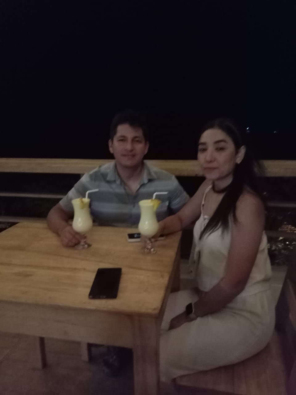 Two people seated at a wooden table with drinks, a smartphone on the table, and a dark background.