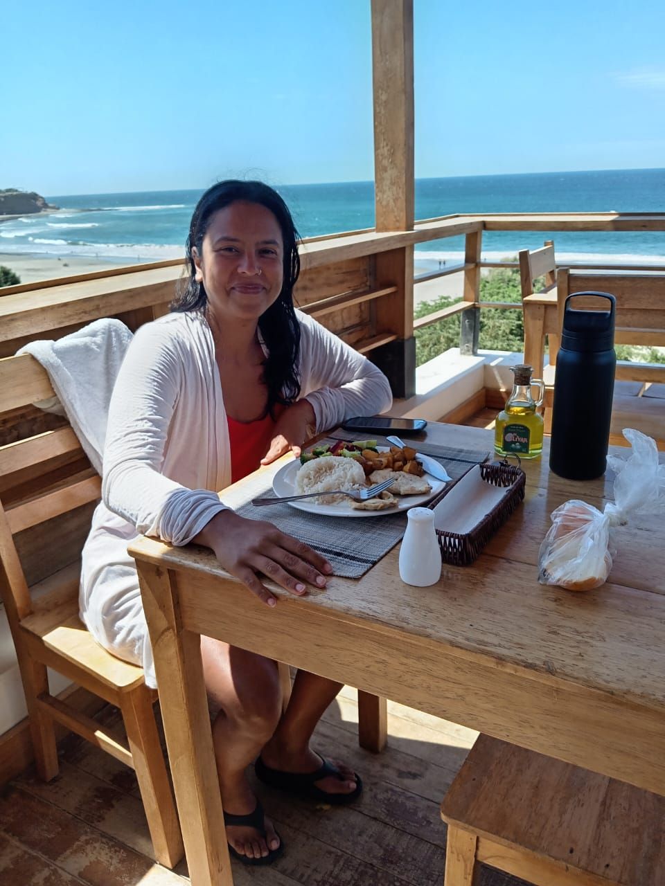Iemand zit aan een houten tafel met eten, met uitzicht op een strand en de oceaan.