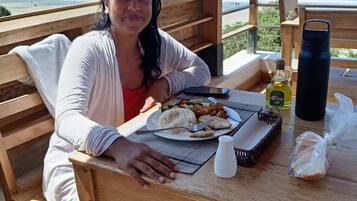 Iemand zit aan een houten tafel met eten, met uitzicht op een strand en de oceaan.