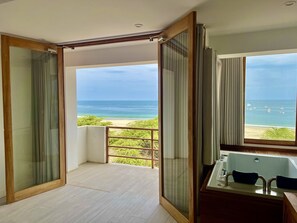 A modern bathroom with a bathtub, a shower, and a large window offering a view of the beach and ocean.