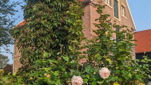 Een gebouw van baksteen met een rode dakbedekking, omringd door weelderige begroeiing en bloeiende planten.