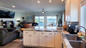 A modern kitchen with white cabinets, a granite countertop, and a large window overlooking a deck. - Tides and Sunshine - Ocean Views w/ Private Pools! (North Topsail Beach)