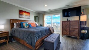 A bedroom with a large bed, a wooden headboard, a nightstand, a bench, a dresser, and a TV mounted on the wall. - Tides and Sunshine - Ocean Views w/ Private Pools! (North Topsail Beach)