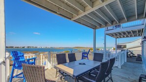 A covered outdoor deck with a table and chairs, overlooking a body of water and distant shoreline. - Good Day Sunshine: Pool +Dock +Ocean Views! (North Topsail Beach)