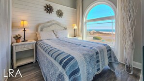 A bedroom with a large bed, two bedside lamps, a decorative wall piece, and a view of the beach through a window. - Good Day Sunshine: Pool +Dock +Ocean Views! (North Topsail Beach)