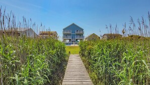 A wooden pathway leading to a blue house with a balcony, surrounded by tall grass. - Good Day Sunshine: Pool +Dock +Ocean Views! (North Topsail Beach)