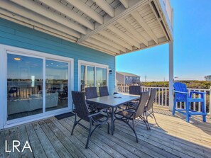 A covered outdoor deck with a table and chairs, a blue chair, and a view of the ocean and distant buildings. - Good Day Sunshine: Pool +Dock +Ocean Views! (North Topsail Beach)