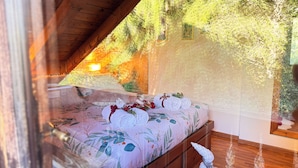 Une chambre avec un plafond en bois, un lit recouvert d’une literie à motifs floraux et une vue sur les arbres par la fenêtre.