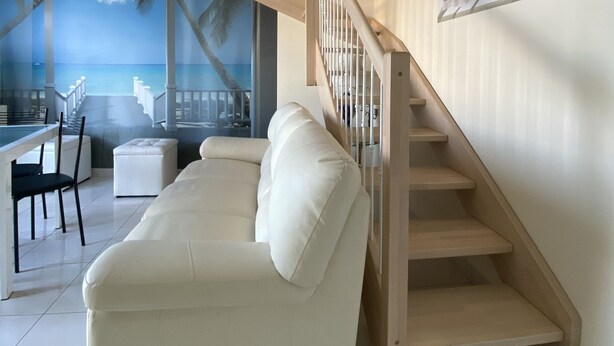 A modern living room with a white sofa, a wooden staircase, and a large window with a view of the sea.