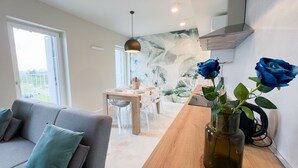 A modern living room with a sofa, a dining area with a table and chairs, and a kitchen area with a stove. There are two blue roses in a vase on the dining table. - Farmont Seafront Acquamarine Suite by CaSa Check (Chioggia)