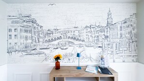 Eine Wandmalerei zeigt eine Stadtansicht mit einer Brücke, ein Schreibtisch mit Blumen in einer Vase, eine Lampe und ein paar Papiere.