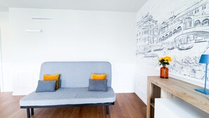 Ein graues Sofa mit zwei Kissen, ein Holztischchen mit einer Blumenvase und eine Wand mit einer Schwarzweißzeichnung.