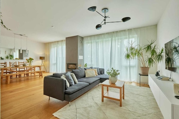 A modern living room with a grey sofa, wooden dining table, and a large window with sheer curtains.