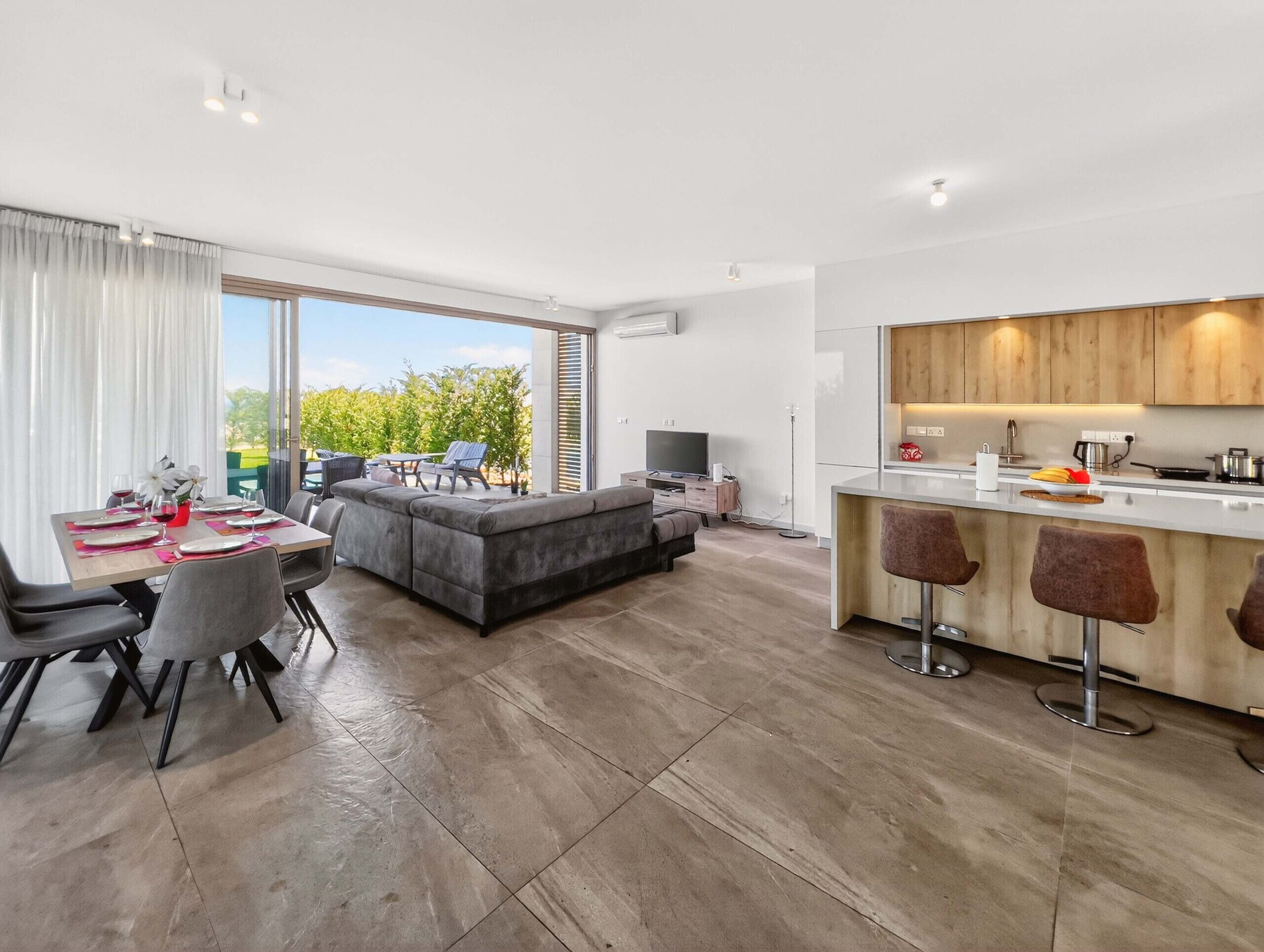 A modern apartment with a dining area, a living room with a sofa, a kitchen with bar stools, and a large sliding glass door to the outdoors.