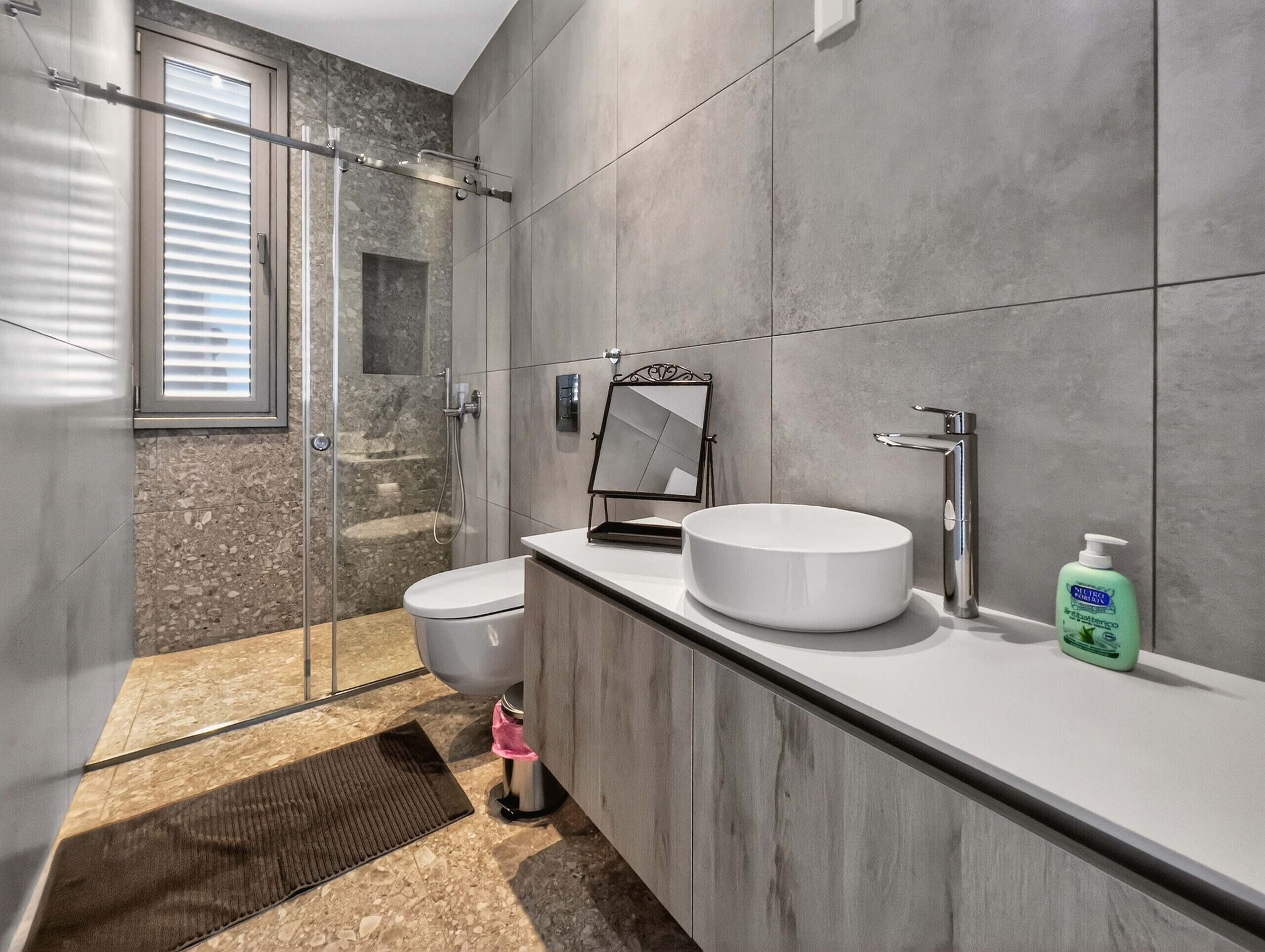 Un baño moderno con cabina de ducha de vidrio, inodoro, lavabo con espejo y una botella de loción verde sobre la encimera.