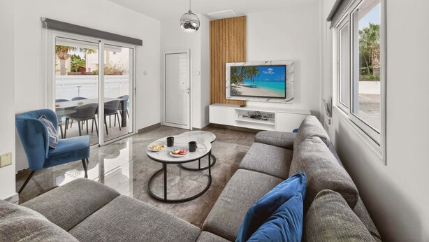 A modern living room with a sofa, a coffee table, a TV on a white cabinet, and a sliding glass door leading to an outdoor area. - Elite Luxury Villa / Private Pool / Privacy (Protaras)