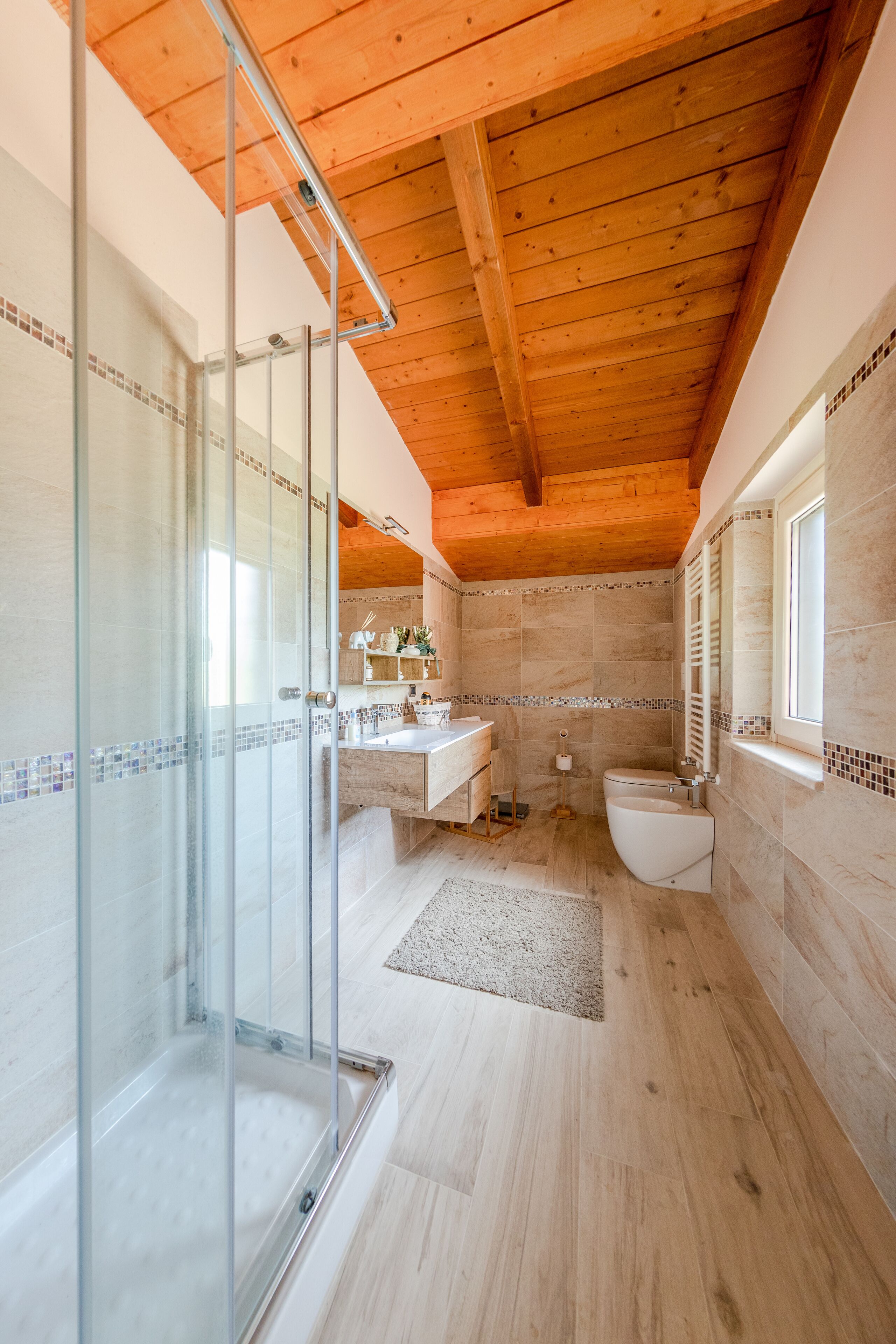 A modern bathroom with a glass shower enclosure, wooden ceiling beams, and a tiled backsplash.