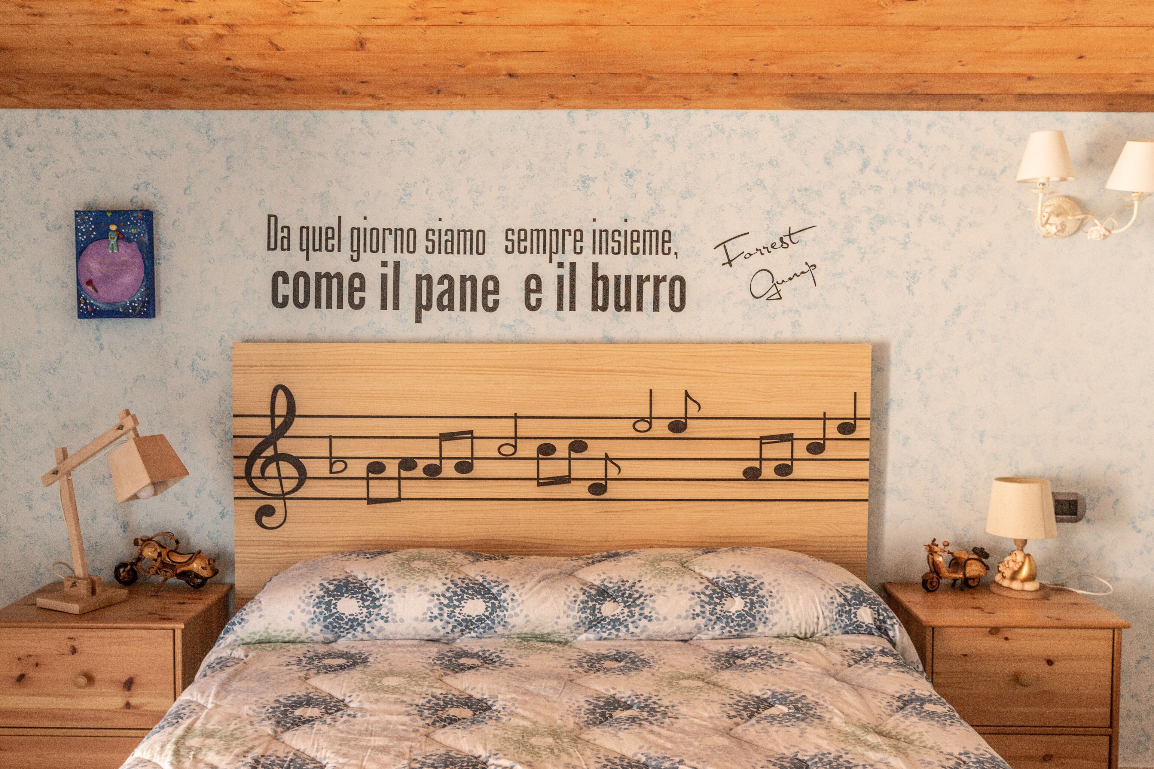 A bedroom with a wooden headboard, a bed with a floral patterned bedspread, a nightstand with a lamp, and a wall with a musical note design and a quote in Italian.