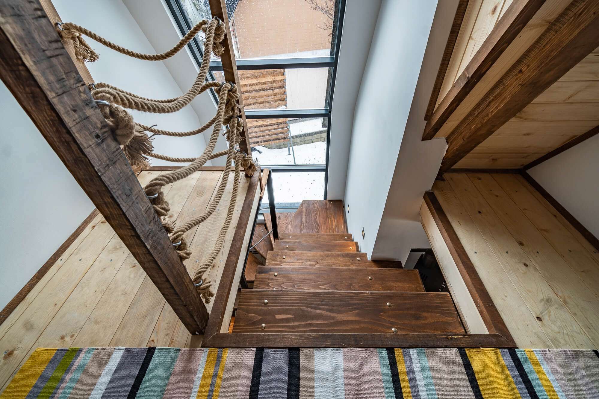 Un escalier en bois, avec une barrière faite de cordes sur le côté gauche, mène à une fenêtre d’où l’on peut admirer les bâtiments environnants.