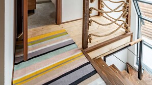 Una escalera de madera lleva a un dormitorio en altillo con tragaluz, alfombra a rayas y una escalera de cuerda.