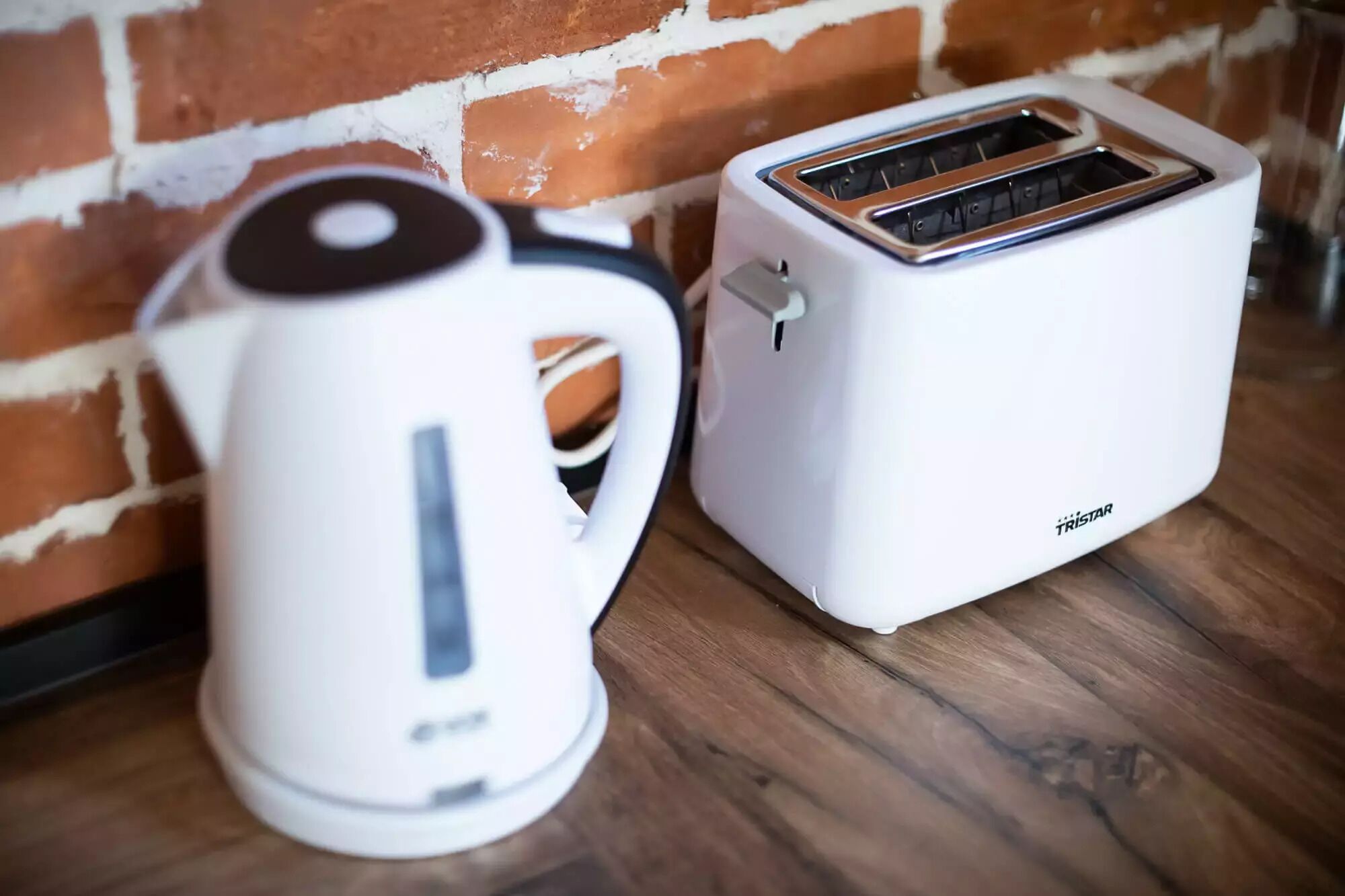 Ein weißer Wasserkocher und ein weißer Toaster auf einer Holztischplatte vor einer Backsteinwand.