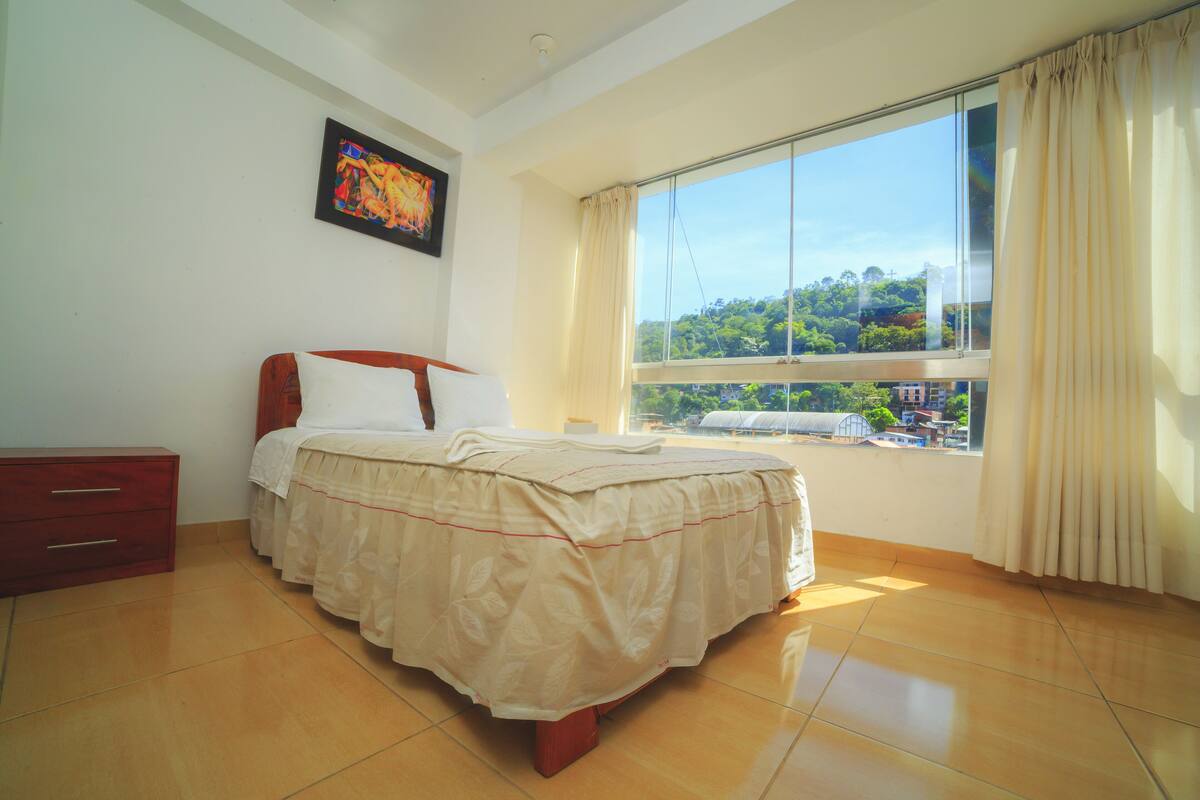 Une chambre à coucher comprenant un lit, une table de chevet, une commode et une grande fenêtre avec des rideaux.