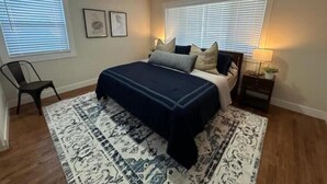 A bedroom with a bed, two windows with blinds, a chair, a nightstand with a lamp, and a patterned rug. - Cozy Mountain Escape (Taylorsville)
