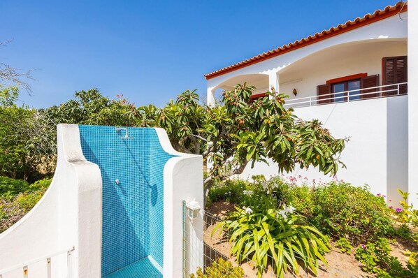 A pool with blue tiles, a white building with a red roof, and various plants and flowers around.
