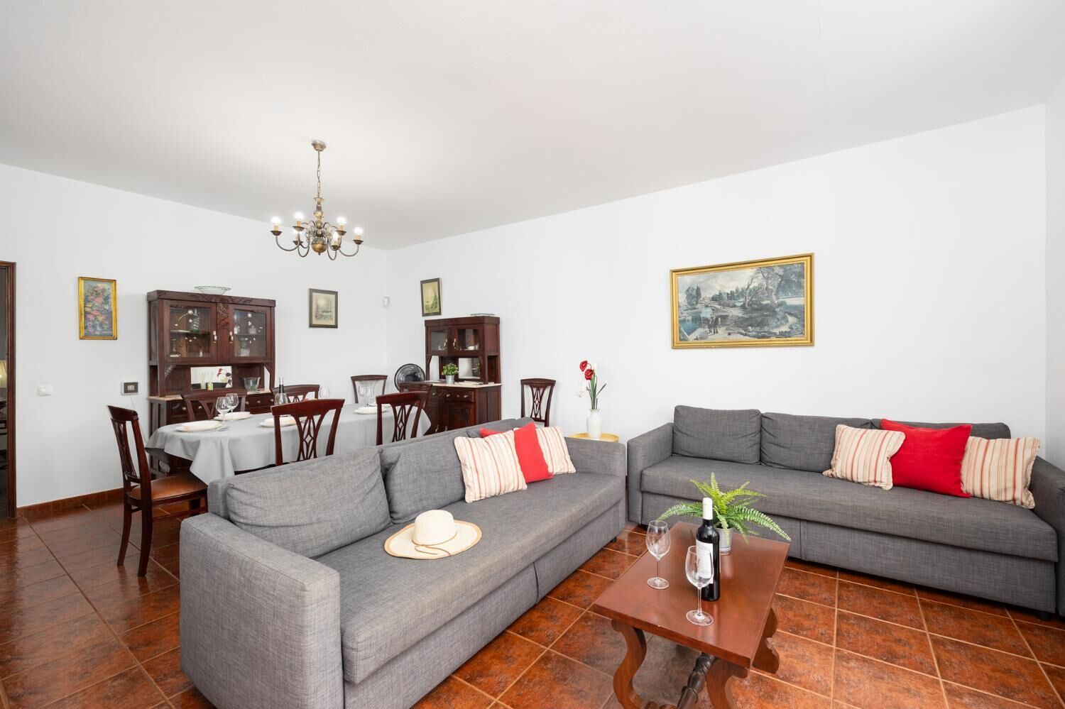 Een woonkamer met een grijze bank, een eettafel met stoelen, een kroonluchter en een schilderij aan de muur.