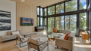 A spacious living room with a fireplace, large windows, and a view of trees. - Boulder Place Lookout At Northstar - TLUXP (Truckee)
