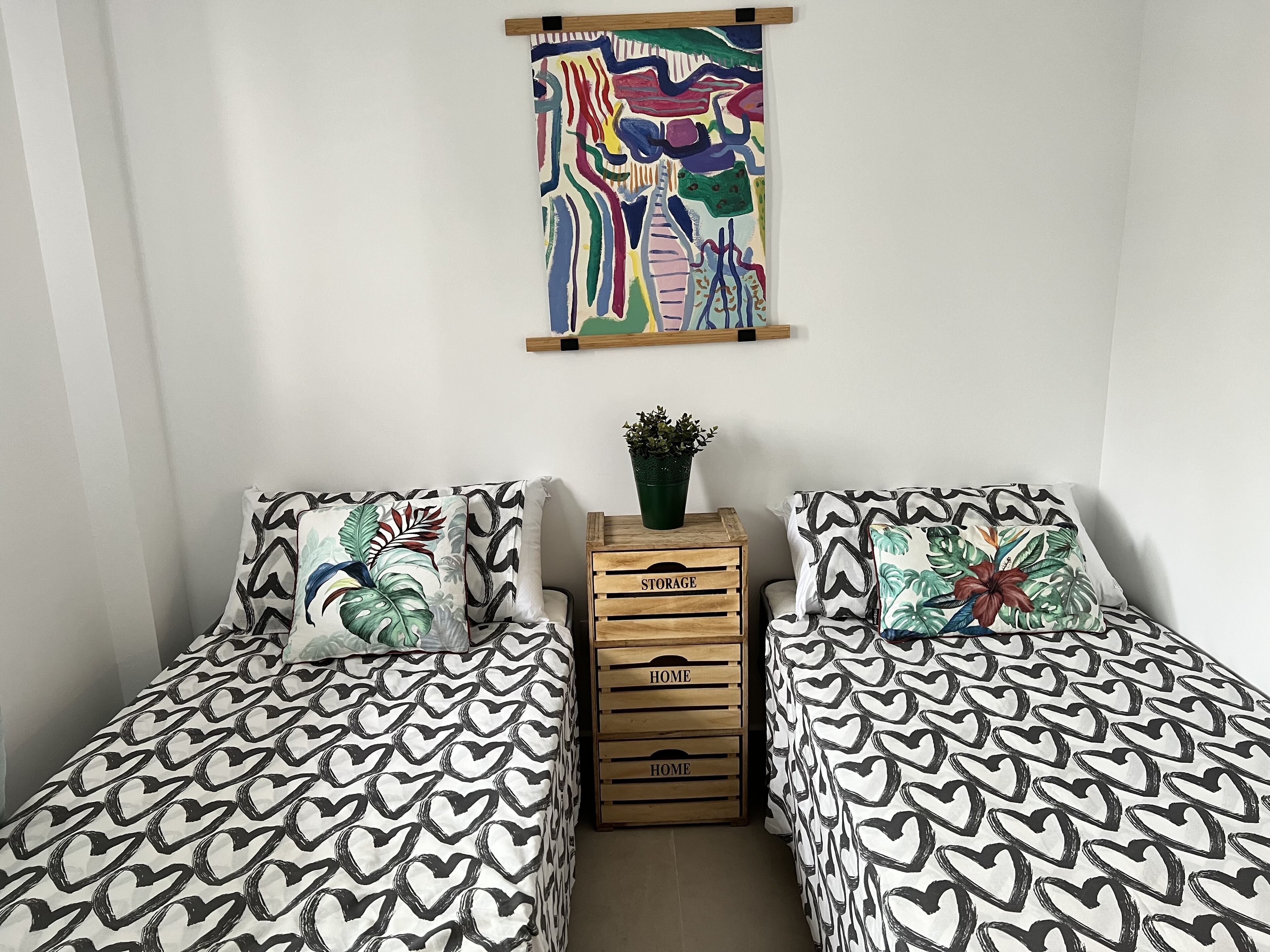 Ein Schlafzimmer mit zwei Betten, jeweils mit einem gemusterten Überwurf, einem an der Wand befestigten abstrakten Gemälde und einem kleinen Topf mit Pflanze auf einer mit 'STORAGE' beschrifteten Holzkiste.
