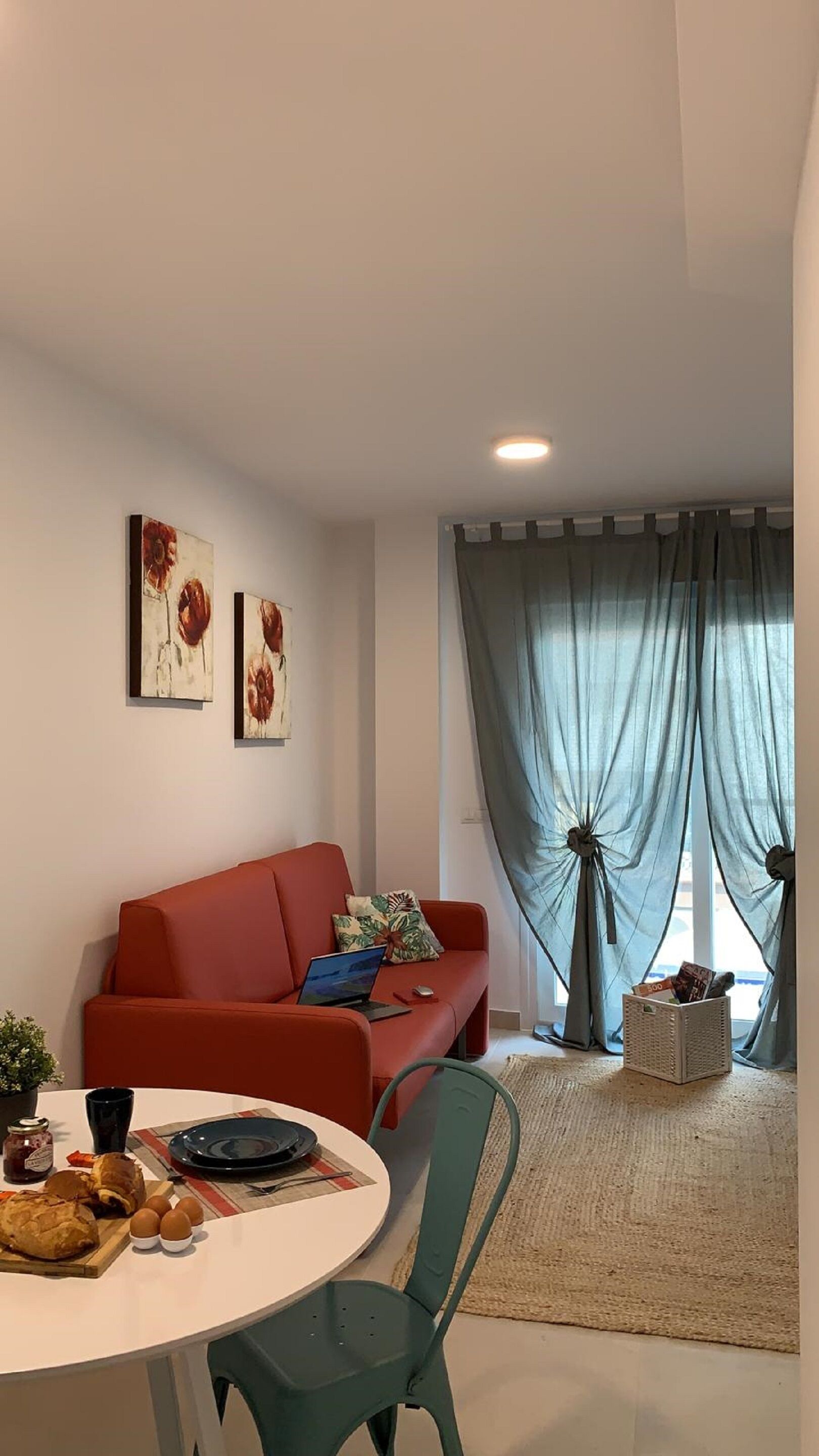 Een moderne woonkamer met een rode bank, een eettafel met stoelen en een groot raam met gordijnen.