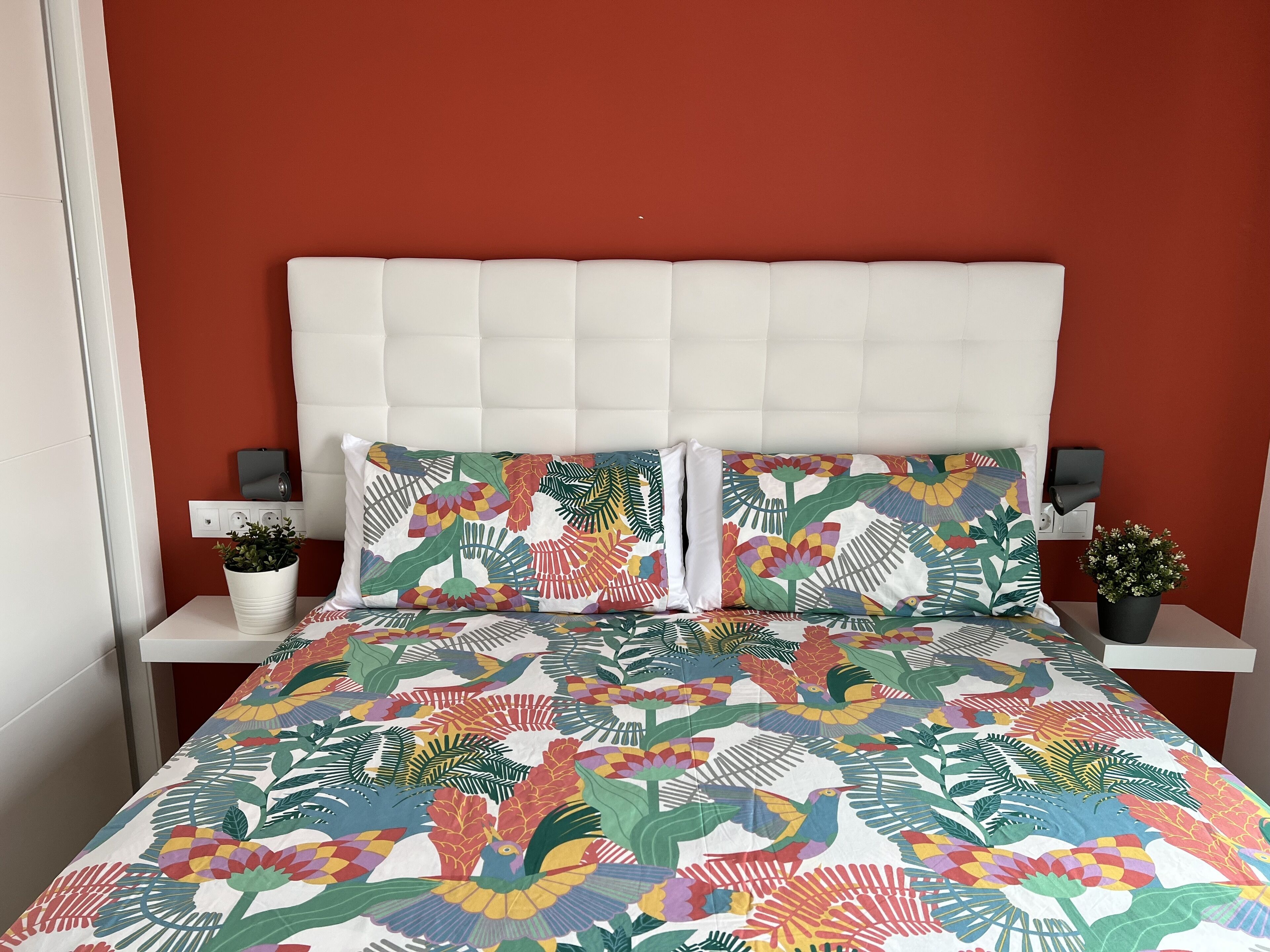 Ein Bett mit farbenfroher Bettdecke mit floralem und Vogelmuster, ein weißer Kopfteil und passende Kissen. Zwei Nachttische mit kleinen Topfpflanzen darauf.