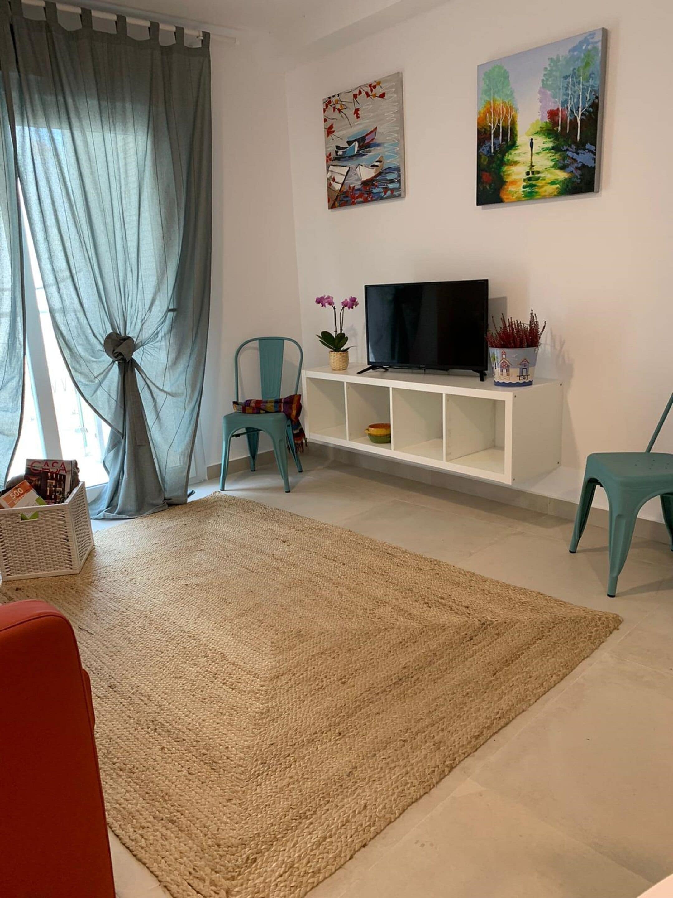 Una sala de estar con un tapete beige grande, un televisor sobre un estante blanco, dos sillones y una ventana con cortinas azules.