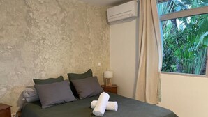 A bedroom with a green bedspread, two pillows, a nightstand with a lamp, and a window with curtains. - Moehani Beach Lodge (Puna'auia)
