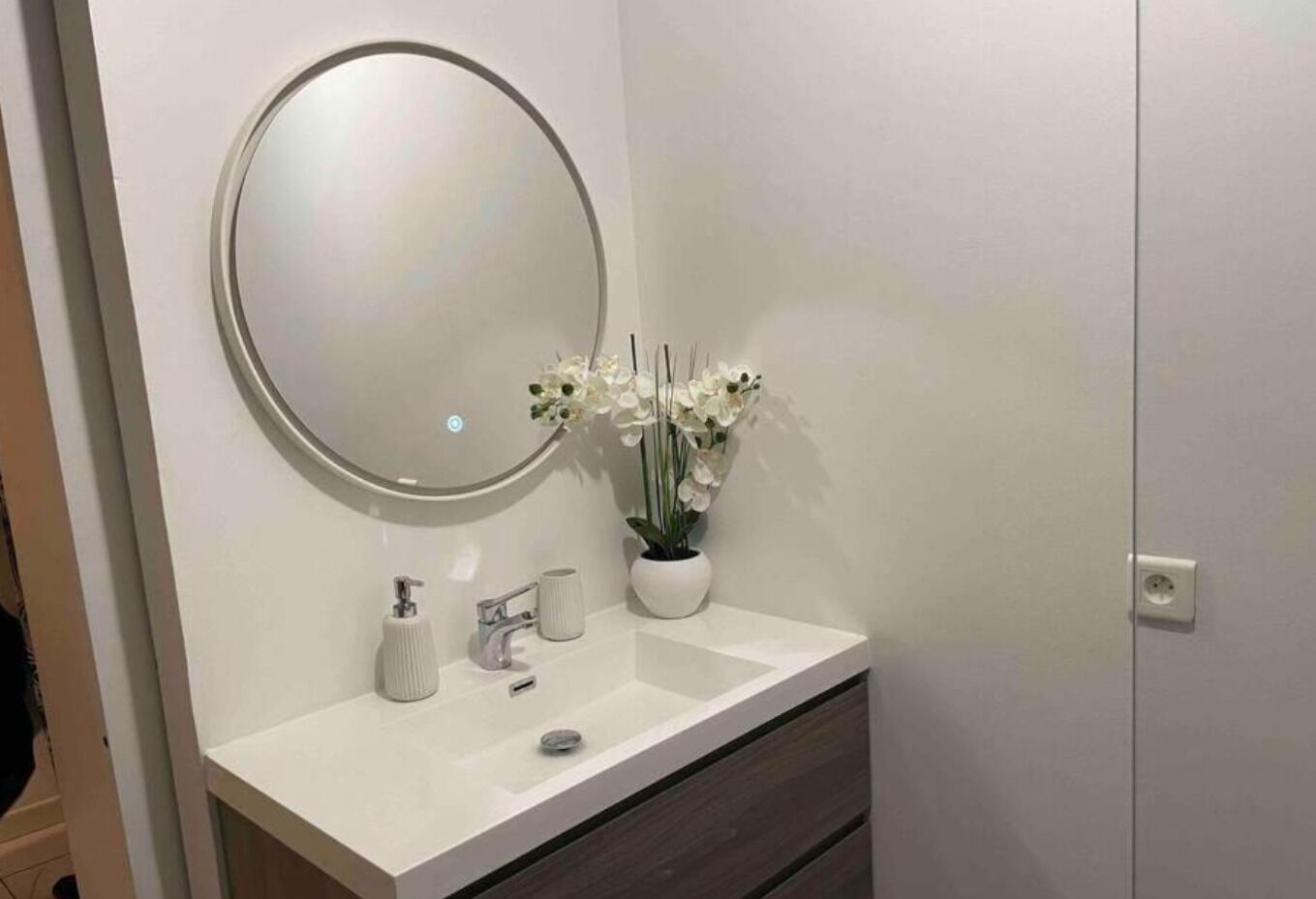 Une salle de bain avec un meuble-lavabo blanc, un miroir rond, un lavabo blanc, un robinet, une petite plante en pot avec des fleurs blanches et un tiroir en bois foncé situé sous le lavabo.