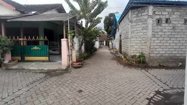 Un sendero pavimentado conduce a una entrada protegida por un portón verde y un edificio blanco con techo azul.