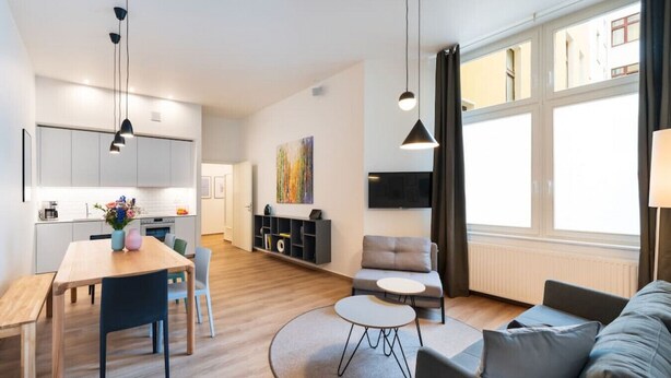 Ett modernt vardagsrum med en soffa, ett soffbord och en matplats med ett bord och stolar. I bakgrunden finns ett kök med skåp och en tavla på väggen.
