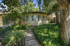A traditional house with a wooden pathway leading to the entrance, surrounded by lush greenery and trees. - "Merced Manor: Luxurious 6BR Home for Ultimate Comfort and Convenience" (Merced)
