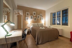 A bedroom with a bed, bedside table, desk with a lamp, wall art, and a window with blinds. - Linas Home - Kalamata Mediterranean Villas (Kalamata)
