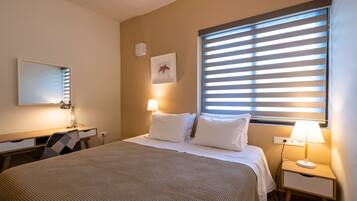 A hotel room with a bed, bedside lamps, a desk with a chair, a mirror, and a window with blinds.