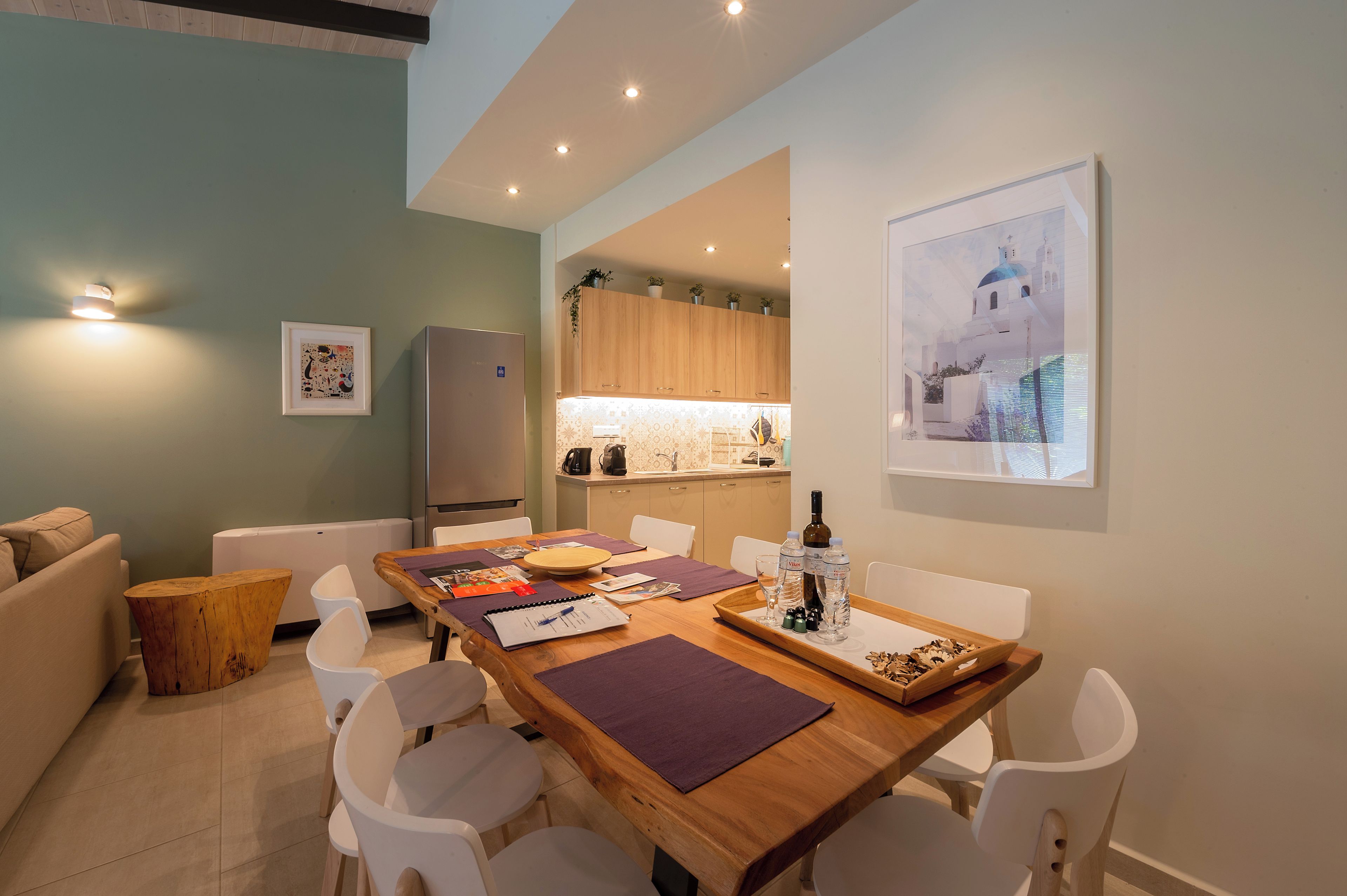 A modern living room with a wooden dining table, white chairs, a small wooden side table, and a sofa. A kitchen is visible in the background with wooden cabinets and a white countertop.