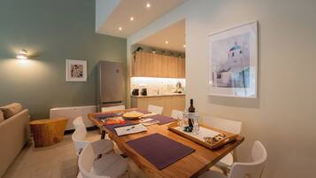 A modern living room with a wooden dining table, white chairs, a small wooden side table, and a sofa. A kitchen is visible in the background with wooden cabinets and a white countertop.